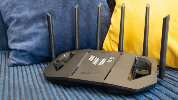 The Asus TUF Gaming BE9400 Wi-Fi 7 router seen from the front, with an angular black body, TUF Gaming logo, vented centre panel and six upright antennas, resting on a blue ribbed cushion.