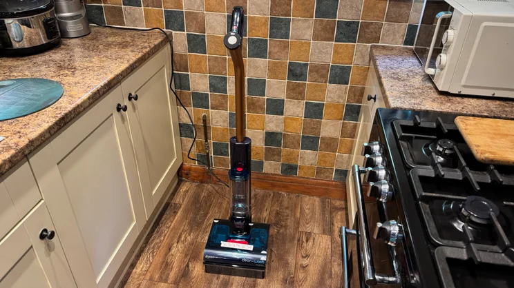 Dyson Clean and Wash Hygiene cordless wet floor cleaner standing upright on wood-effect flooring in a kitchen with mosaic wall tiles, cream cabinets and a gas hob