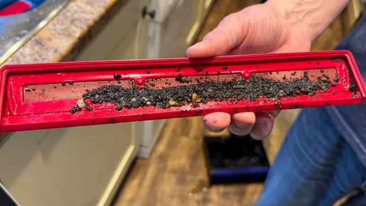 Hand holding the red debris tray from the Dyson Clean and Wash Hygiene, full of dark grit, dirt and small debris over the mesh filter