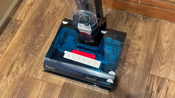 Close-up from above of the Dyson Clean and Wash Hygiene cleaner head with its transparent blue cover, red debris tray and a setup instruction sticker