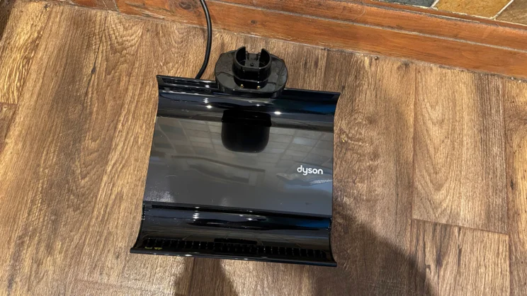 Empty black charging and self-clean dock tray for the Dyson Clean and Wash Hygiene, with its cable and charging connector, on a wood-effect floor