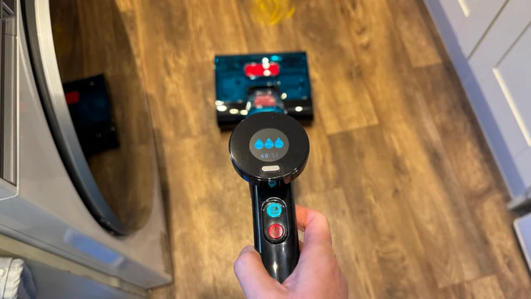 Handheld view of the Dyson Clean and Wash Hygiene display showing three water droplets and battery status time, with the cleaner head on the floor ahead