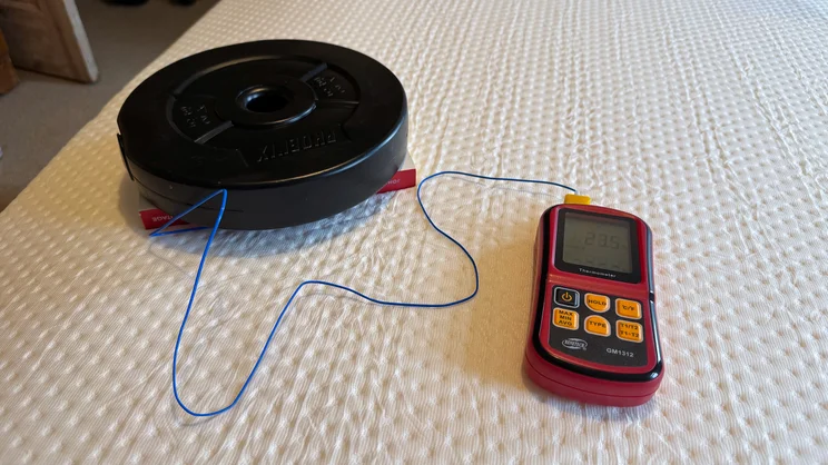 A digital thermocouple thermometer being used to measure temperature retention on the surface of the Emma Original mattress. A 2.5kg weight plate and paperback book are holding down the temperature probe to the surface of the mattress