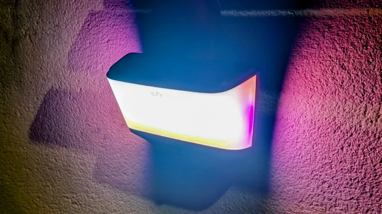 Eufy S4 4K Solar Wall Light glowing at night, casting pink, purple and yellow colored light across a textured stucco wall.