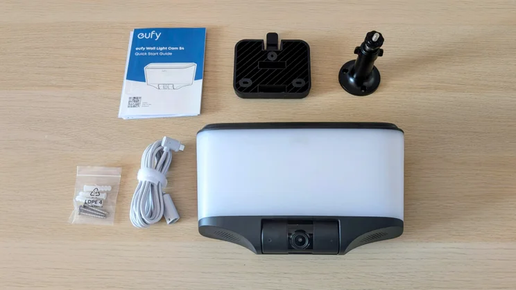 Eufy S4 4K Solar Wall Light unboxed on a wooden table beside its quick start guide, wall mounting plate, black adjustable bracket, white extension cable and screws with wall plugs.