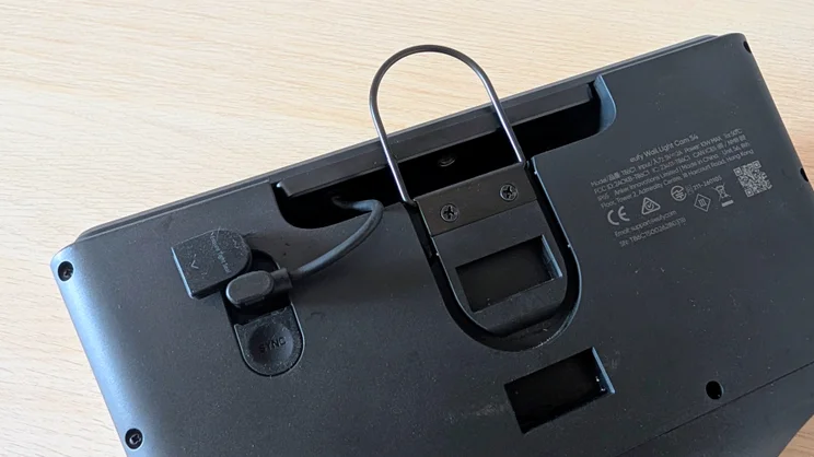 Close-up of the back of the Eufy S4 4K Solar Wall Light showing the wire hanging loop, the SYNC button under a rubber cover, the charging port with cable attached and the regulatory label.