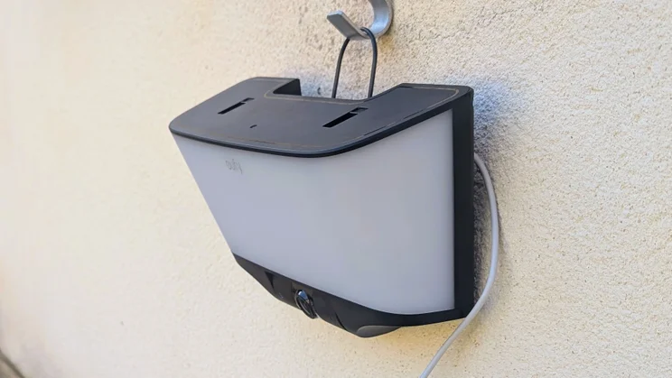 Eufy S4 4K Solar Wall Light hanging from a metal hook on an outdoor wall by its wire loop, with the solar panel removed from the top and a white cable running down the side.