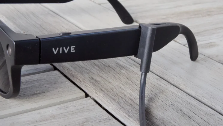 Close-up of the HTC Vive Eagle smartglasses arm with the Vive logo and a button, and a charging cable clipped onto the left arm