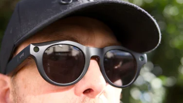 Close-up of the HTC Vive Eagle smartglasses being worn by a man wearing a black cap, showing the corner camera, the round lenses and the Vive logo on the arm