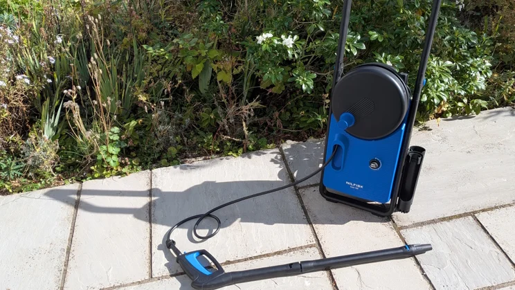 Nilfisk Core 150 PowerControl pressure washer standing on paving beside a planted border, with the hose running out to the trigger gun and lance.