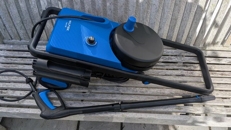 Nilfisk Core 150 PowerControl lying on its back on a weathered wooden bench, with the black hose reel, folding handle, trigger gun and lance beside it.