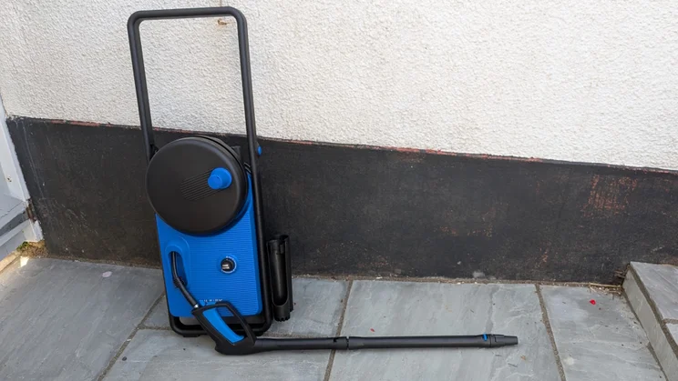 Nilfisk Core 150 PowerControl leaning against a rendered wall on grey paving, with its folding handle raised and the trigger gun and lance laid out in front.