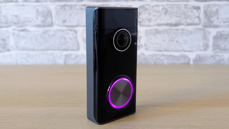 Tapo D260 Pro doorbell standing upright on a wooden surface against white brick wallpaper, with a purple light ring around its metallic button.
