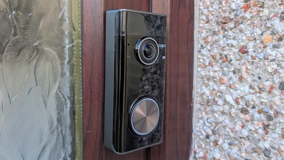 Tapo D260 Pro doorbell mounted on a dark wood-effect door frame, between patterned obscured glass and a pebbledash wall, seen from a side angle.