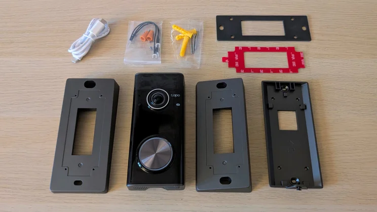 Tapo D260 Pro doorbell laid out on a wooden table with its box contents: USB-C cable, wiring connectors, screws and wall plugs, a gasket, a 3M VHB adhesive pad and three mounting brackets