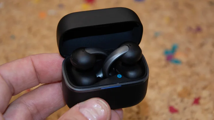 A hand holding the opened matte black charging case of the Anker Soundcore AeroClip 2, displaying the clip-on earbuds nested inside with an illuminated blue indicator light