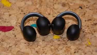 Side profile of both Anker Soundcore AeroClip 2 clip-on earbuds resting side by side on a textured cork surface, displaying the ergonomic C-shaped earhook curve