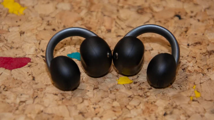 Side profile of both Anker Soundcore AeroClip 2 clip-on earbuds resting side by side on a textured cork surface, displaying the ergonomic C-shaped earhook curve