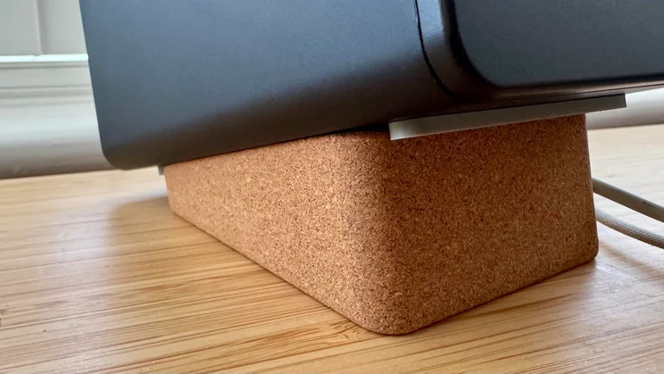A side close-up showing the bottom of a black Creative XF1 desktop speaker resting on an angled, 15-degree cork wedge stand on a wooden desk