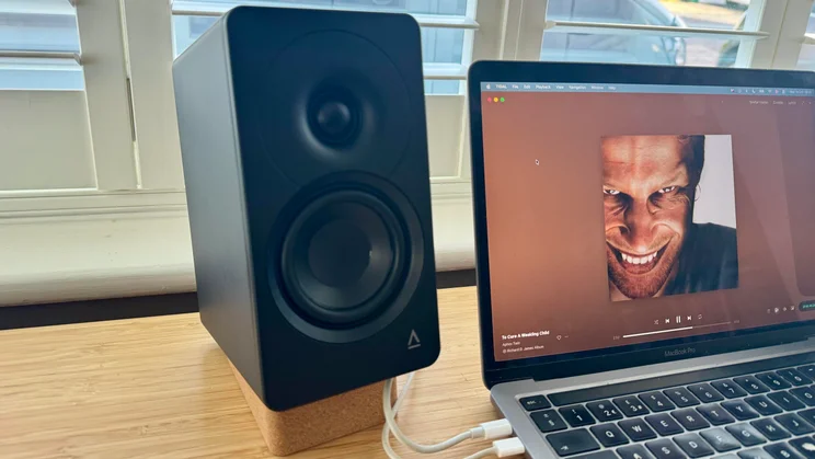 Angled front-side view of the left Creative XF1 desktop speaker positioned on a wooden desk next to a laptop
