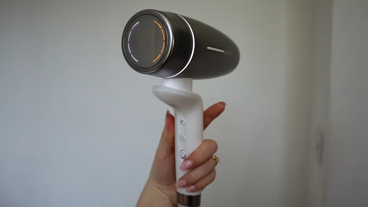 A hand holding up the GHD Speed Ionic hair dryer, facing away from the camera, against a white wall background