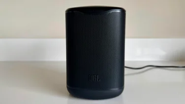 Front view of the black JBL Cove M1 wireless speaker standing upright on a counter, showing the wrap-around speaker grille with the JBL logo at the base