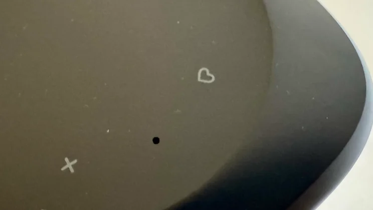 Extreme close-up of the top panel of the JBL Cove M1 wireless speaker, focusing on a microphone pinhole beside a heart-shaped preset button