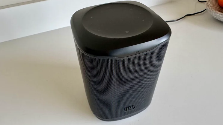 Angled high view of the black JBL Cove M1 wireless speaker resting on a white worktop, showing both the top control surface and front mesh grille