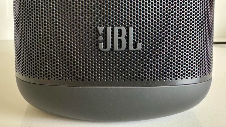 Close-up of the lower front of the JBL Cove M1 wireless speaker, showing the black metal perforated grille and embossed JBL brand logo above the base