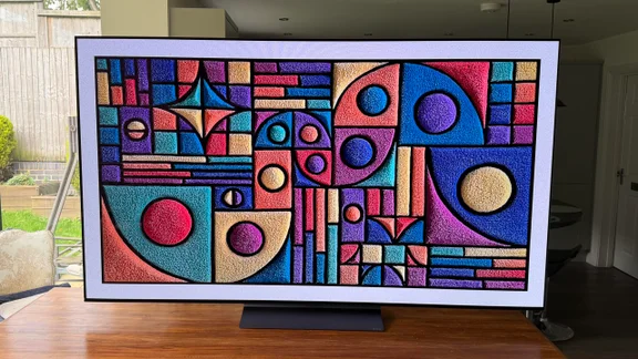 Direct front view of the 77in LG C6 OLED TV with Primary RGB Tandem OLED technology resting on a wooden tabletop, displaying a vibrant, highly detailed geometric carpet pattern art piece with rich colours and deep blacks