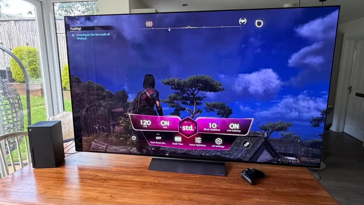 Wide shot of an Xbox Series X console and controller next to the 77in LG C6 OLED TV with Primary RGB Tandem OLED technology, displaying a gaming screen with the active Game Optimiser menu overlay