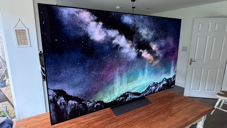 Angled side view of the 77in LG C6 OLED TV sitting on a wooden tabletop, showcasing its ultra-thin bezel and vivid display with Primary RGB Tandem OLED panel technology rendering a colourful galaxy and starry night sky