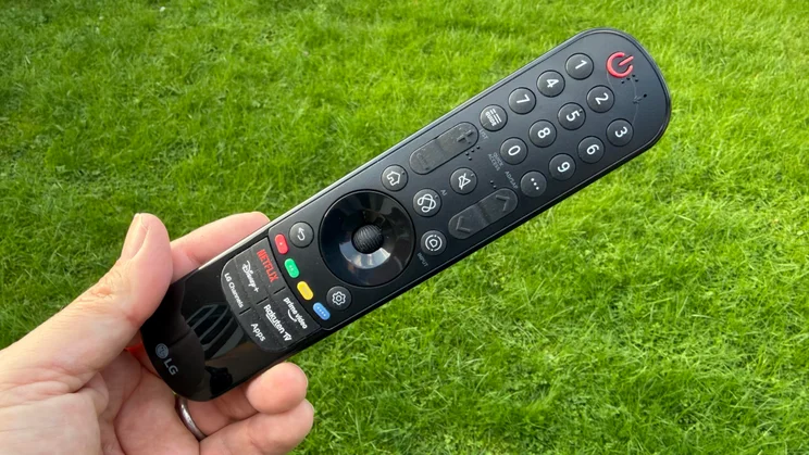Close-up of a hand holding the black LG Magic Remote for the 77in LG C6 OLED TV featuring Primary RGB Tandem OLED technology, featuring dedicated streaming shortcut buttons and a central scroll wheel against a green grass background