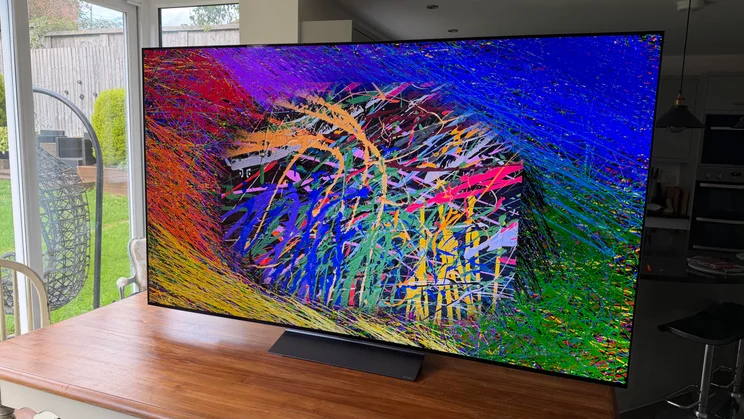 Right-angled view of the 77in LG C6 OLED television resting on a wooden table near a glass sliding door, highlighting the bright colour output and deep contrast of its Primary RGB Tandem OLED display showing complex, multi-coloured abstract line artwork