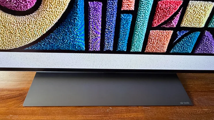 Close-up view of the dark metallic tabletop pedestal stand supporting the 77in LG C6 OLED TV with Primary RGB Tandem OLED technology, showing the subtle "LG OLED" branding etched on the right corner of the base