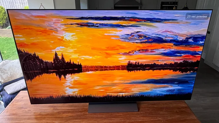 Wide front-facing view of the 77in LG C6 OLED TV resting on a wooden tabletop, showcasing the high contrast and colour range of its Primary RGB Tandem OLED screen displaying a bright orange and yellow sunset landscape painting