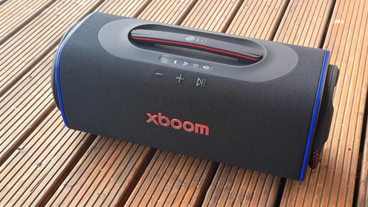 An angled view of the black LG xboom Blast portable Bluetooth speaker resting on a wooden deck, featuring illuminated blue LED light rings on its side passive radiators and visible red "XBOOM" branding on the front