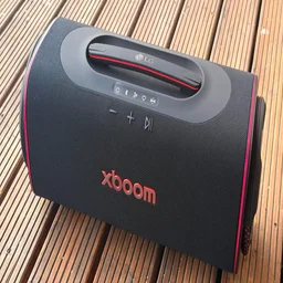 An elevated side-angle view of the LG xboom Blast portable speaker on a wooden surface, highlighting its woven black fabric exterior, integrated top carrying handle, physical control buttons, and red edge lighting