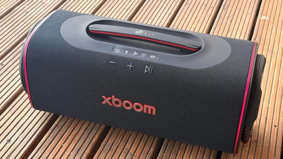 An elevated side-angle view of the LG xboom Blast portable speaker on a wooden surface, highlighting its woven black fabric exterior, integrated top carrying handle, physical control buttons, and red edge lighting