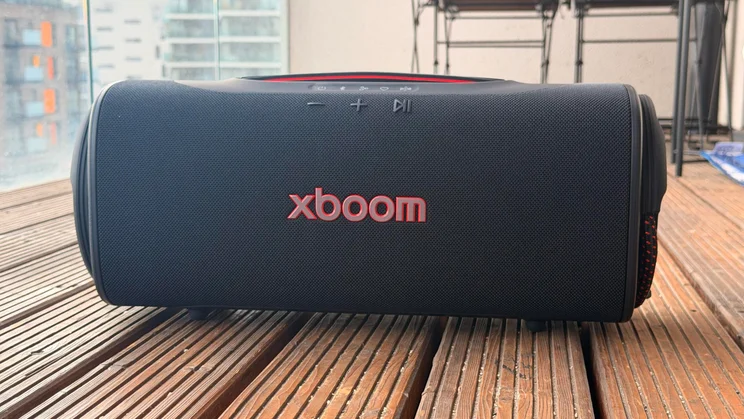 A direct front-facing shot of the LG xboom Blast portable speaker positioned outdoors on wooden decking, displaying the red "XBOOM" logo on the mesh grill and top volume/playback controls