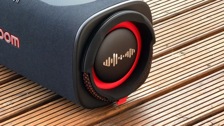 A detailed close-up of one end of the LG xboom Blast speaker, focusing on the circular passive radiator membrane with a red surround, metallic audio waveform icon, and woven side strap loop.