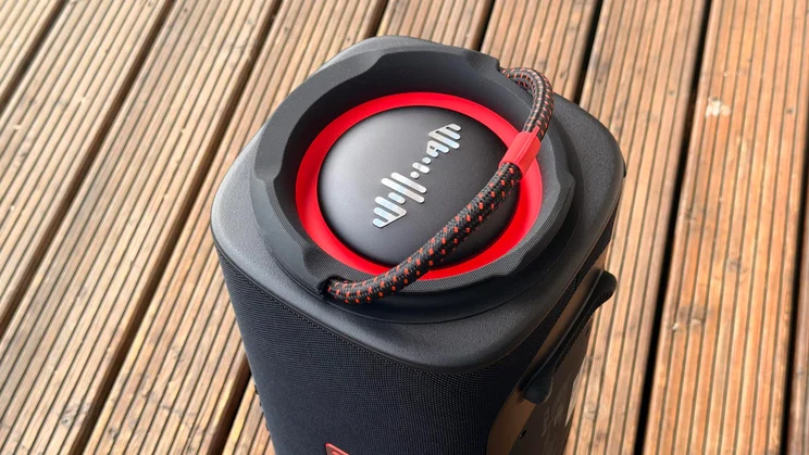 A close-up top-down angle of the side passive radiator on the LG xboom Blast speaker, showing its red accent trim ring, central waveform graphic logo, and attached braided carrying strap