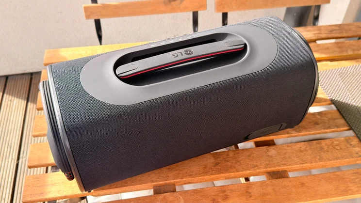 A rear perspective of the LG xboom Blast speaker resting on a wooden slat table, emphasising the durable top carrying handle with red lining and an embossed LG logo