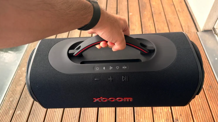 A person's hand lifting the black LG xboom Blast portable speaker by its built-in top handle over outdoor wooden decking, showcasing its portability and physical control panel
