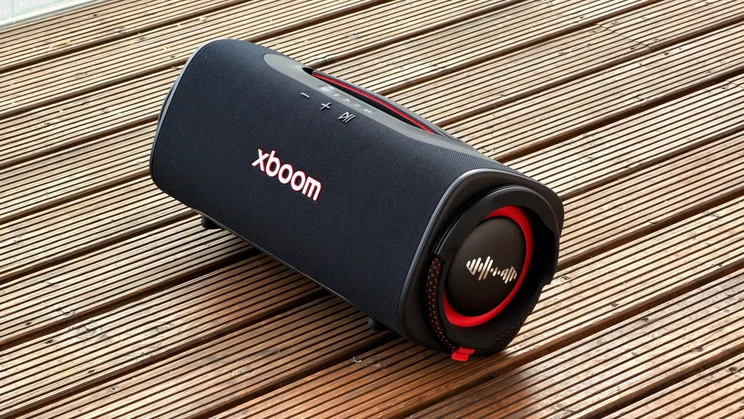 An angled front view of the black LG xboom Blast portable speaker sitting on a wooden deck, showing the front mesh grill, illuminated red "XBOOM" logo, top control panel, and the side passive radiator with a red accent ring