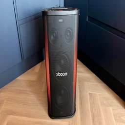 A front view of the upright LG xboom Stage 501 party speaker displaying warm orange-red lighting along its sides on a wooden floor