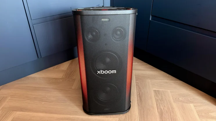 A front view of the upright LG xboom Stage 501 party speaker displaying warm orange-red lighting along its sides on a wooden floor