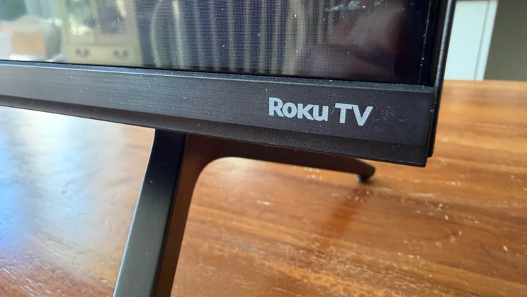 Close-up view of the lower-right bezel on the Sharp HJ6725KB showing the printed "Roku TV" branding above a stand leg
