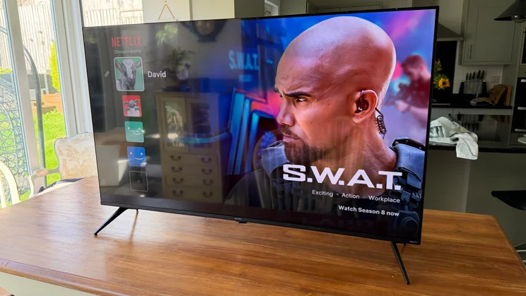 Front right perspective view of the Sharp HJ6725KB Roku TV resting on a wooden table, displaying the Netflix user selection screen