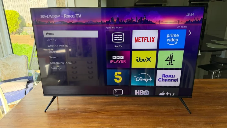 Front view of the Sharp HJ6725KB Roku TV on a wooden table, displaying the Roku OS home screen with streaming apps like Netflix, Prime Video, BBC iPlayer, and Disney+